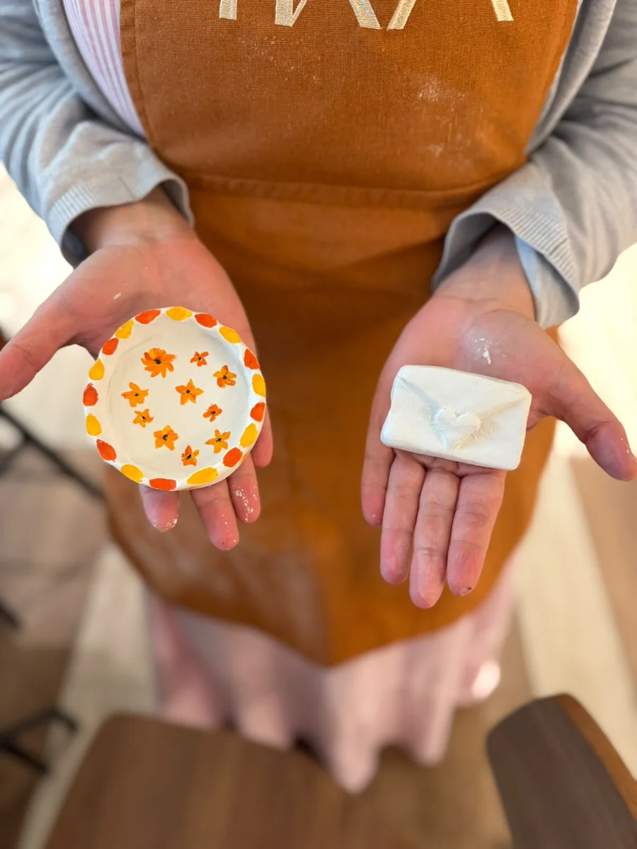Mãos apresentam uma peça redonda com flores laranja e amarelas e outra branca em forma de envelope.