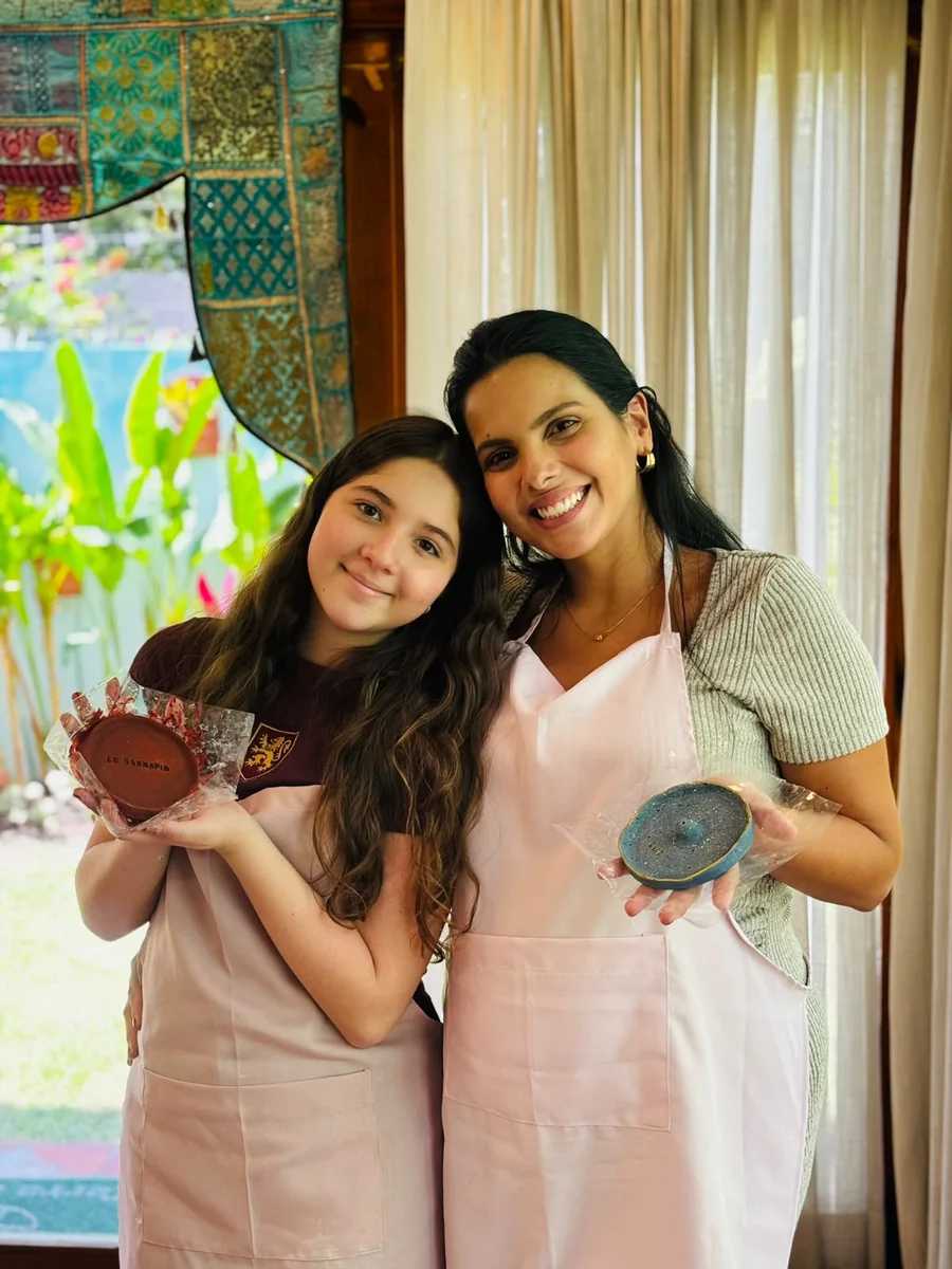 Duas participantes de avental rosa posam juntas mostrando suas peças prontas.